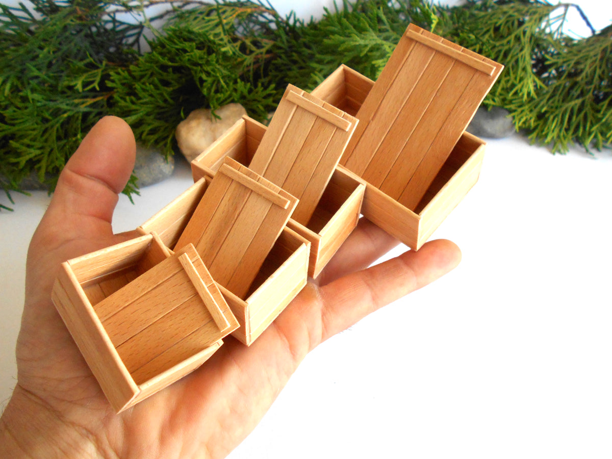 A hand holding several miniature light wood boxes, shown close up against a white background with green foliage at the top.
