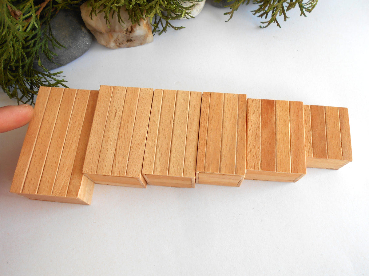 A set of natural wood miniature boxes  arranged in a staggered row on a white surface, with a finger pointing to the leftmost block and green foliage in the background.