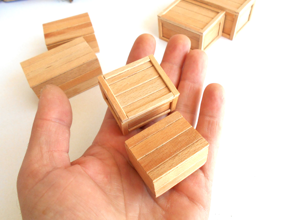 A hand holding three small wooden crate-shaped blocks, with additional similar blocks scattered in the background on a plain white surface.