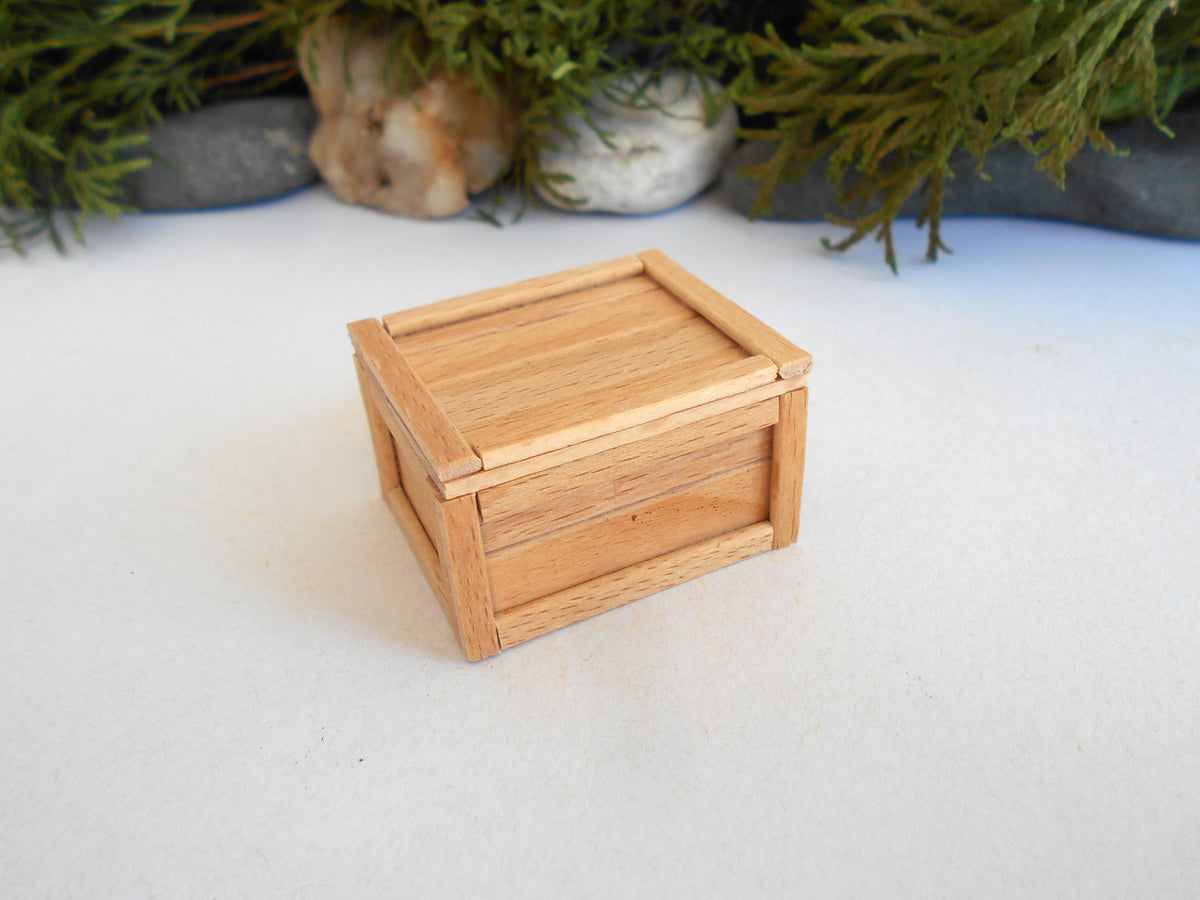 A small handmade wooden box with a hinged lid, made from light natural wood, set on a white surface with a blurred background of green foliage and stones.