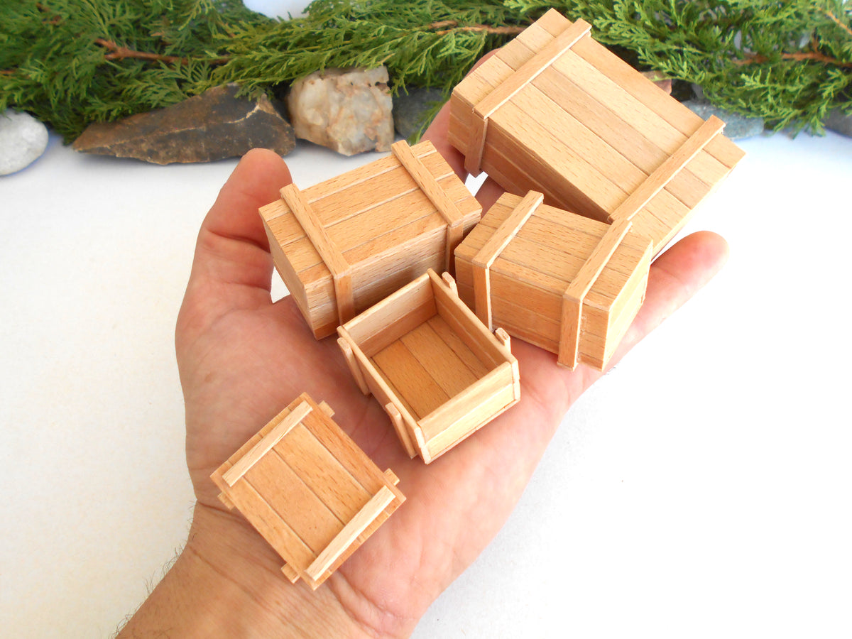 A hand holding several miniature light wood crates of different sizes, with green foliage and small rocks in the background.