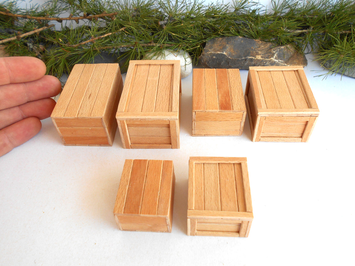 Six miniature wooden crates in different sizes arranged on a white surface, with a hand on the left for scale and pine branches in the background.