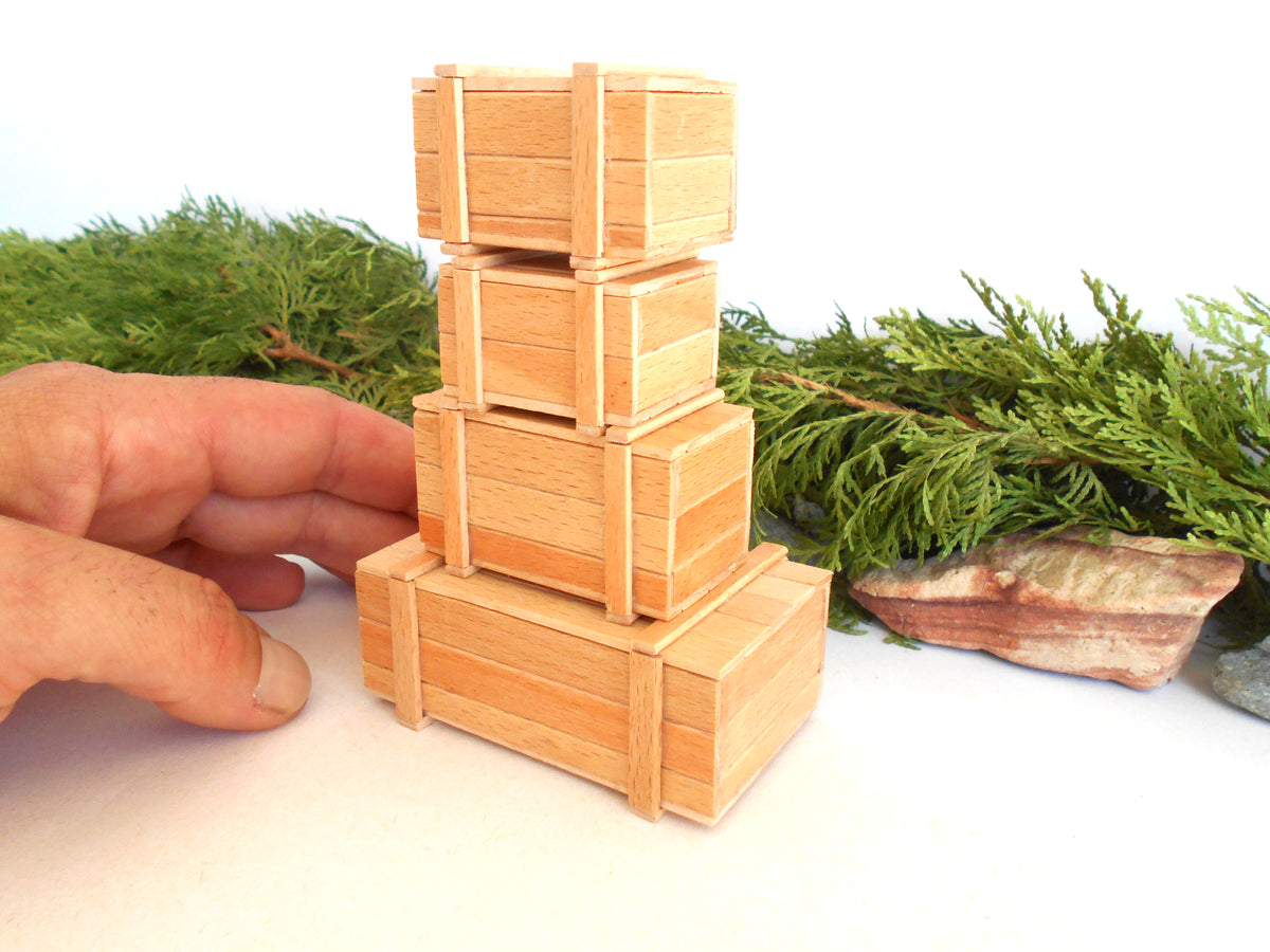 A hand is holding a miniature stack of four small wooden crates, likely made from 3D-printed resin or wood, set against a white background with green foliage props.