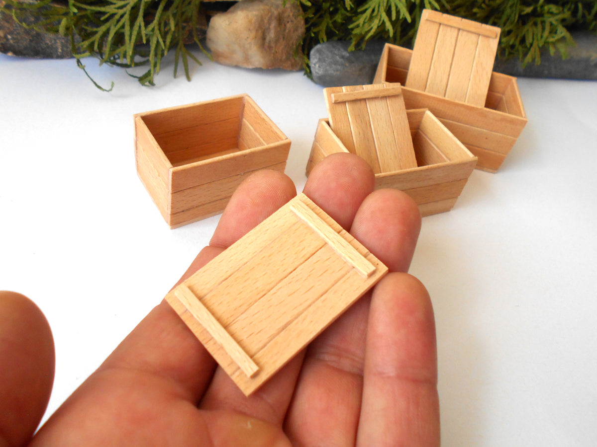 A close-up of a hand holding a miniature light wood crate, with several other tiny wooden crates arranged on a white surface in the background.