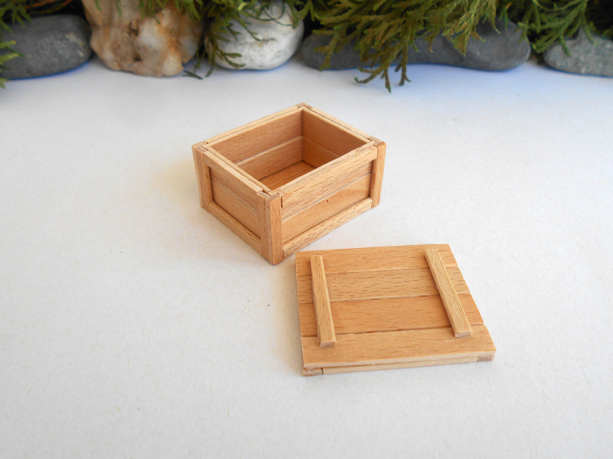 A miniature light wood crate box with its lid placed beside it on a white surface, with small rocks and greenery in the background.