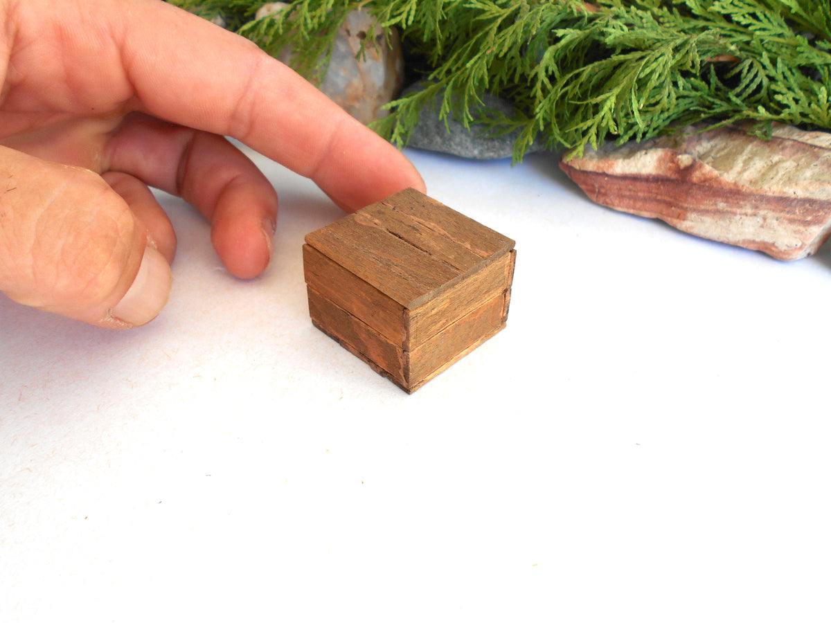 A miniature handcrafted wooden box with a rustic brown finish, held by a person's fingers to demonstrate its tiny scale, set against a white background with decorative greenery.