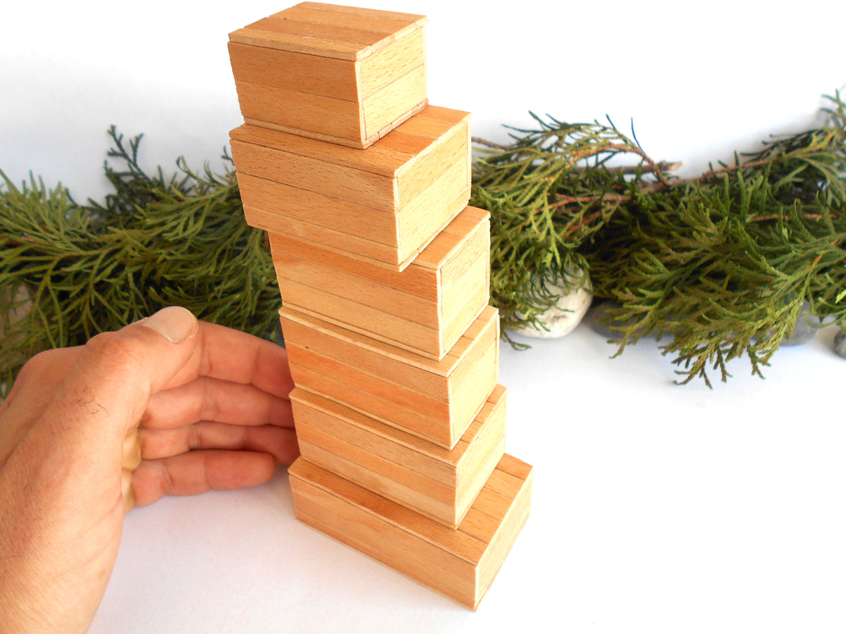 A hand is balancing a stack of natural light wood miniature boxes on a white surface, with green foliage in the background.