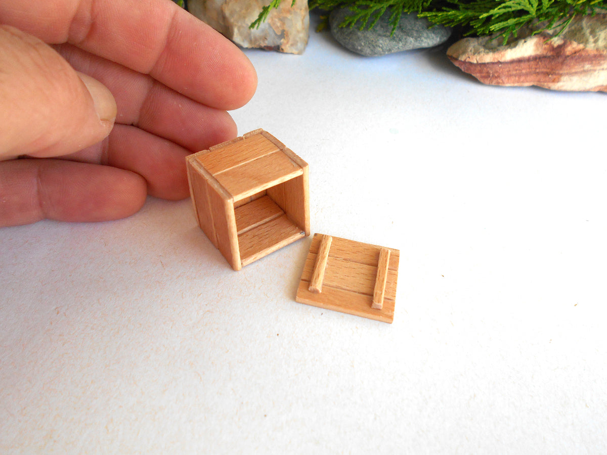A miniature wooden crate or box accessory shown on a white surface, with a human hand holding it for scale and small rocks in the background.