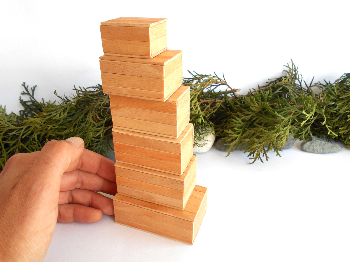 A hand is holding a stack of six small, light-colored wooden miniature boxes arranged in a staggered tower, with green foliage and small stones in the background against a white surface.