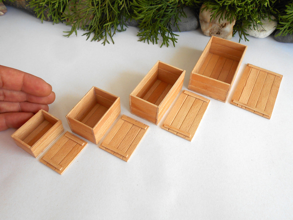 Six miniature light wood boxes and matching lids in various sizes, displayed on a white surface with a hand holding one of the smaller boxes for scale, and green foliage in the background.