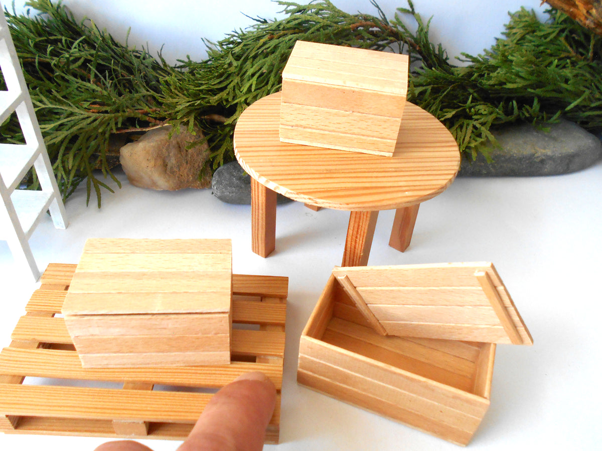 A miniature dollhouse-scale wooden furniture set featuring light natural wood boxes, a round table, and pallet-style bases, displayed on a white surface with green foliage in the background.