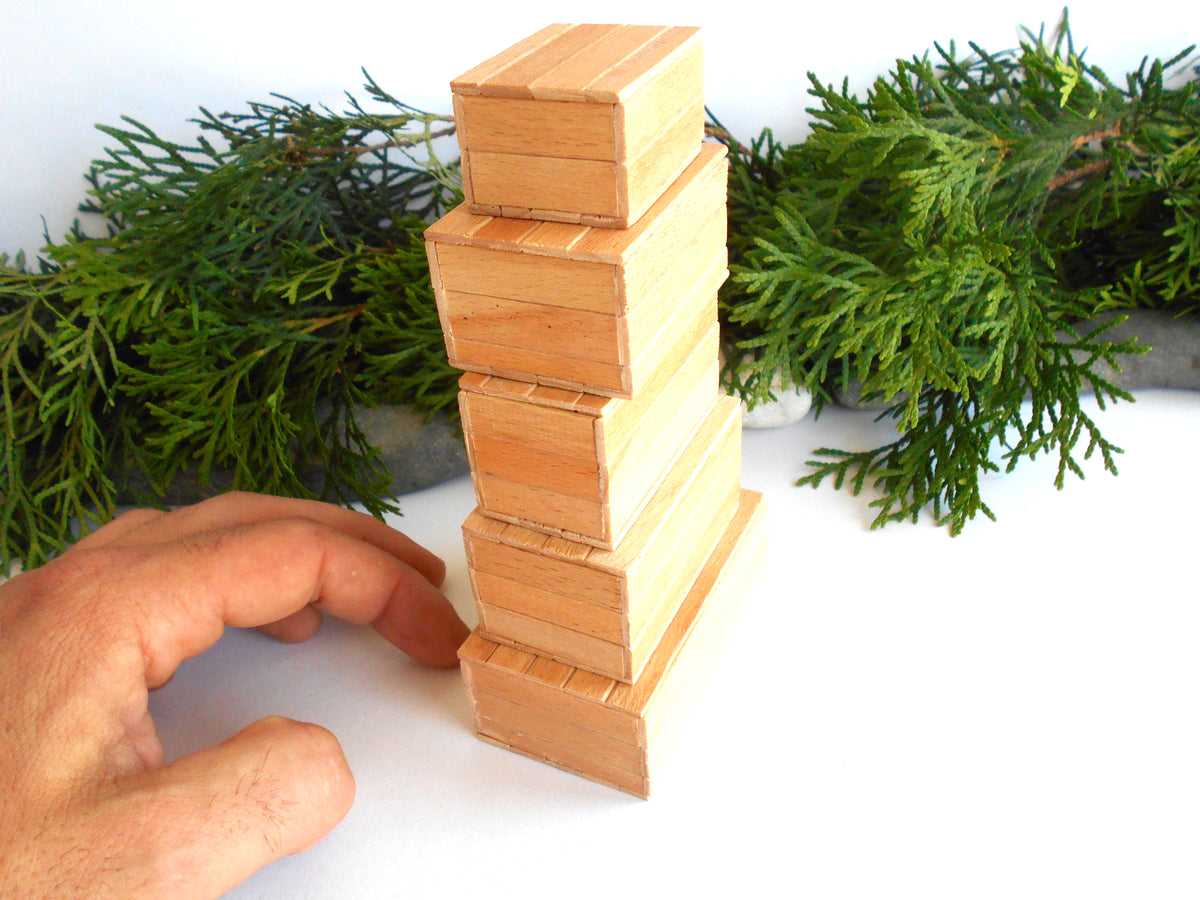 A hand is balancing a small wooden block tower on a white surface, with green foliage in the background.