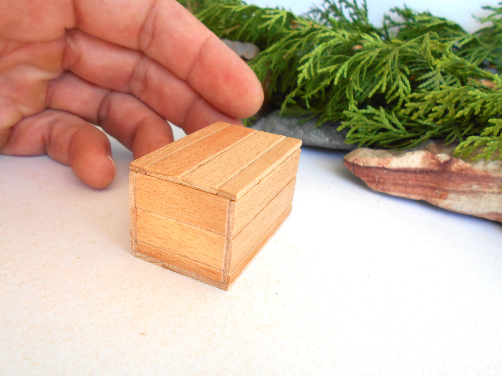 A miniature light brown wooden box with a slatted lid, held by a person's hand to show its small scale, with green foliage in the background.