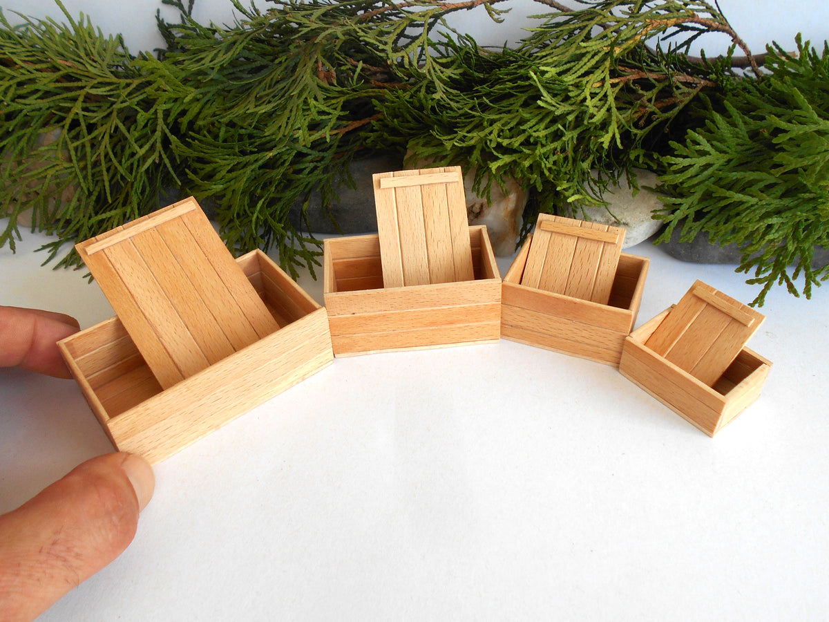 A set of miniature light wood boxes arranged in a semi-circle on a white surface, with a hand holding the leftmost chair and green foliage in the background.