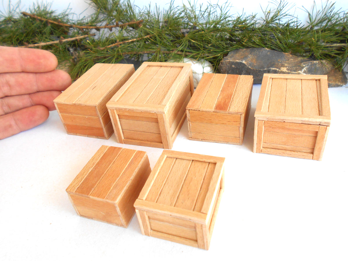 Six miniature light wood crate models arranged on a white surface, with a hand visible on the left for scale and pine branches in the background.