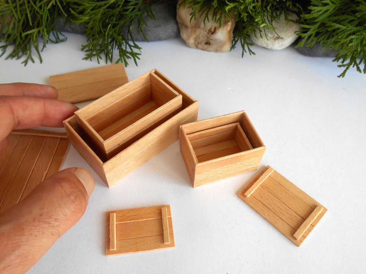 A hand is holding a miniature wooden box with a sliding lid, surrounded by several smaller matching wooden boxes and trays on a white surface with decorative greenery in the background.