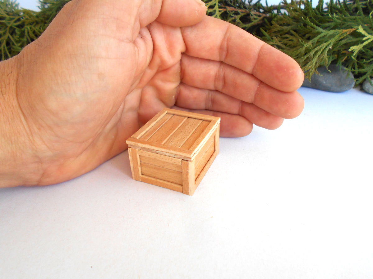 A miniature light wood crate dollhouse piece shown next to a human hand for scale, with a white surface and green foliage in the background.