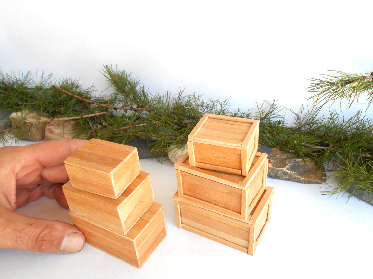 A hand holds a small stack of three light natural wood boxes next to another stack of three matching boxes on a white surface with pine branches in the background.