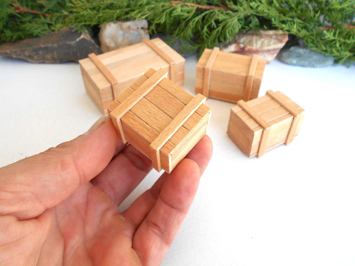 A hand holding a miniature wooden crate, with four other miniature wooden crates of different sizes arranged on a white surface in the background.