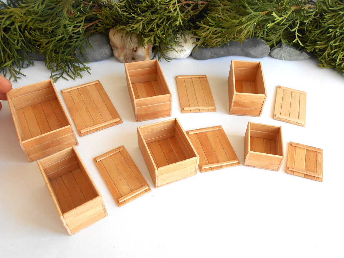 A set of miniature light brown wooden crates and boxes, including both open-top and closed-top styles, arranged on a white surface with green foliage and small stones in the background.