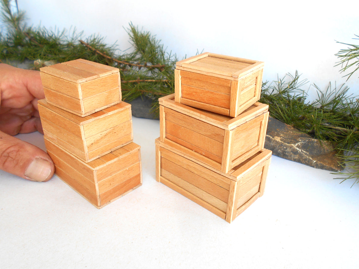 A set of miniature light wood toy boxes, including two small stacked boxes being held by a hand and two larger stacked boxes, displayed on a white surface with a pine garland in the background.