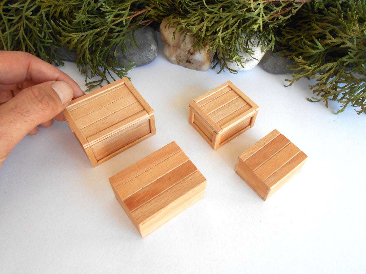 Four miniature light wood crates of different sizes arranged on a white surface with green foliage and stones in the background. A hand is visible on the left holding one of the crates.