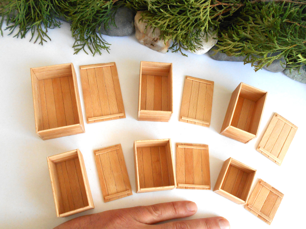 A set of miniature light wood crates and boxes arranged on a white surface, with a hand visible at the bottom for scale and green foliage in the background.