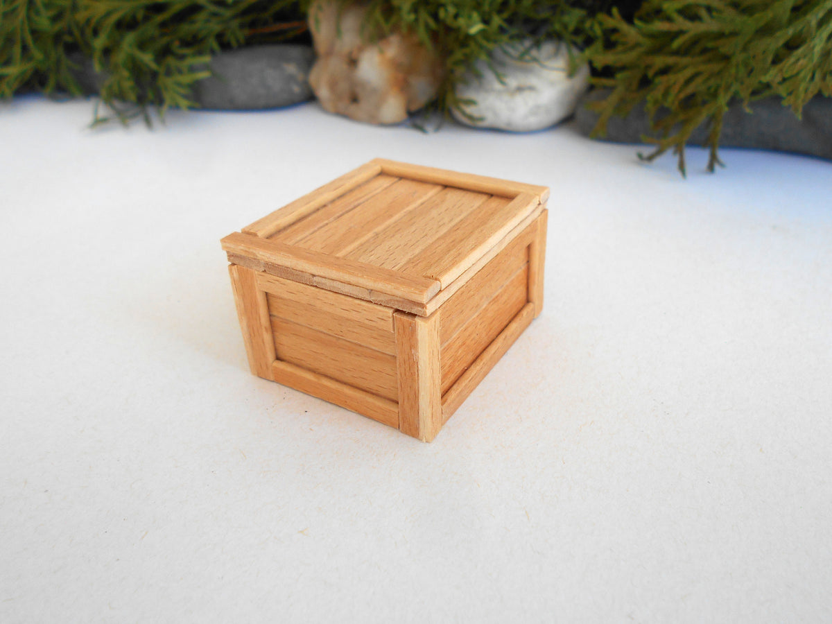 A miniature light brown wooden crate box with a slatted lid, photographed on a white surface with blurred green foliage in the background.