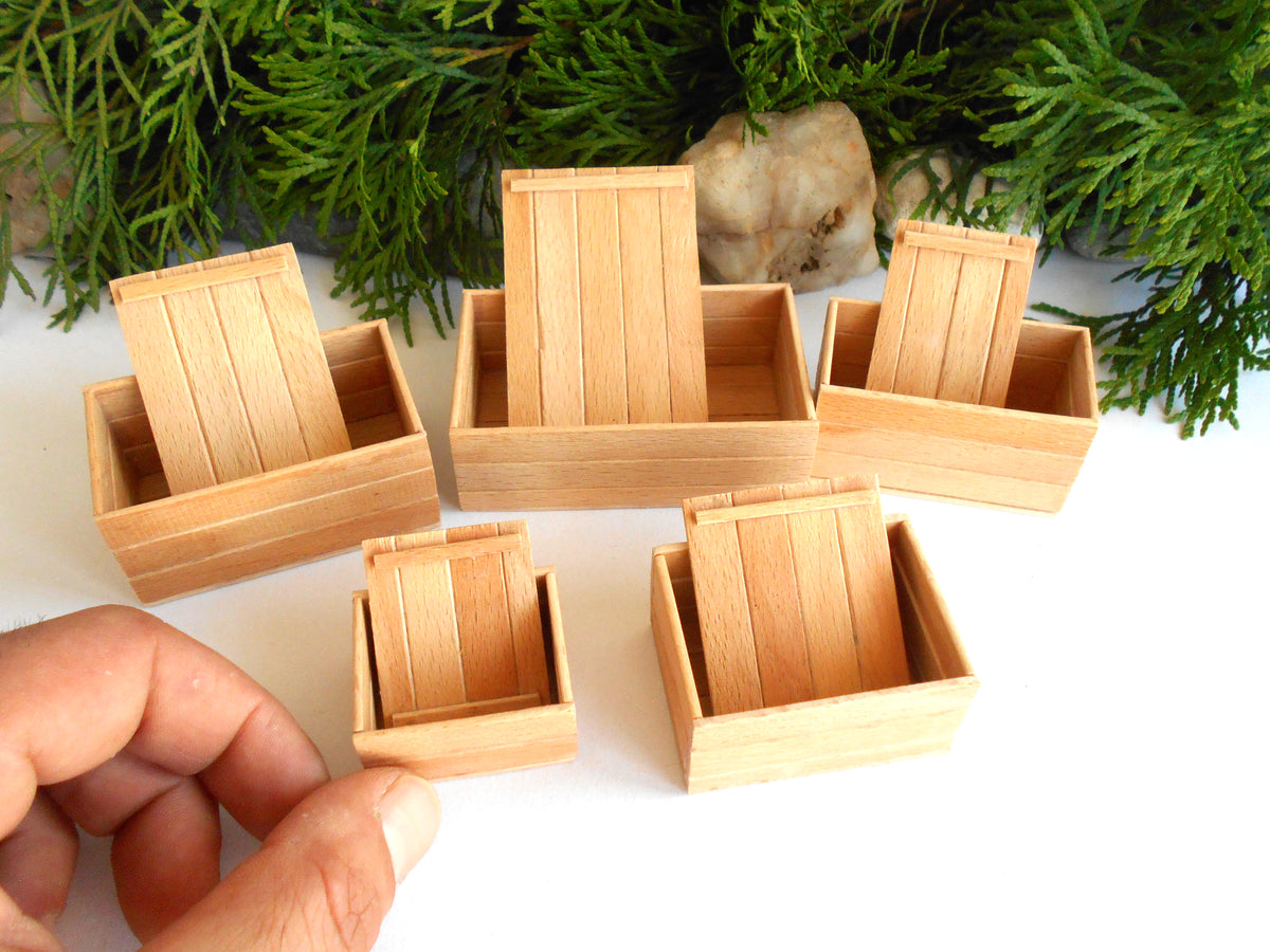 Six miniature light wood boxes of different sizes arranged on a white surface with green foliage in the background. A human hand is visible in the lower-left corner holding one of the small boxes.