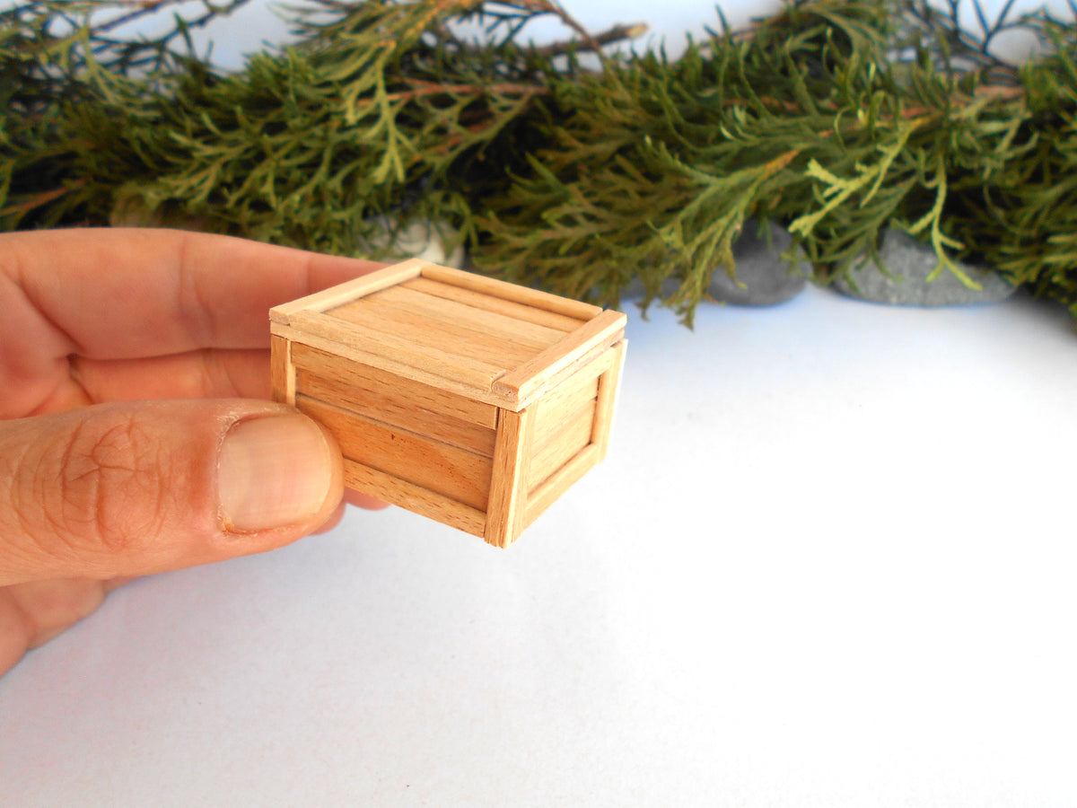 A miniature light wood crate or box being held by a person's hand against a white background with blurred green foliage.