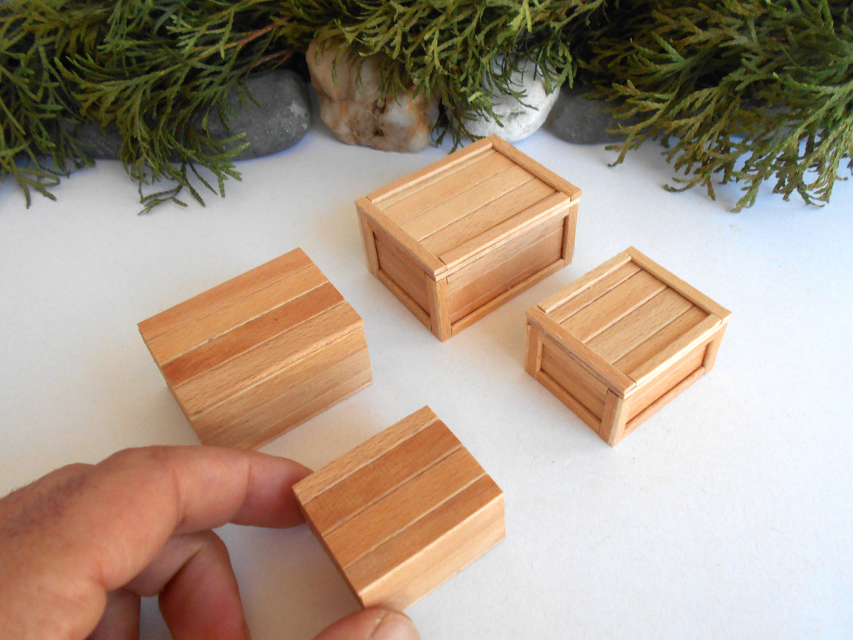 Four small wooden crates or boxes made of light natural wood, with one being held by a hand in the foreground. The boxes are arranged on a white surface with green foliage and stones in the background.