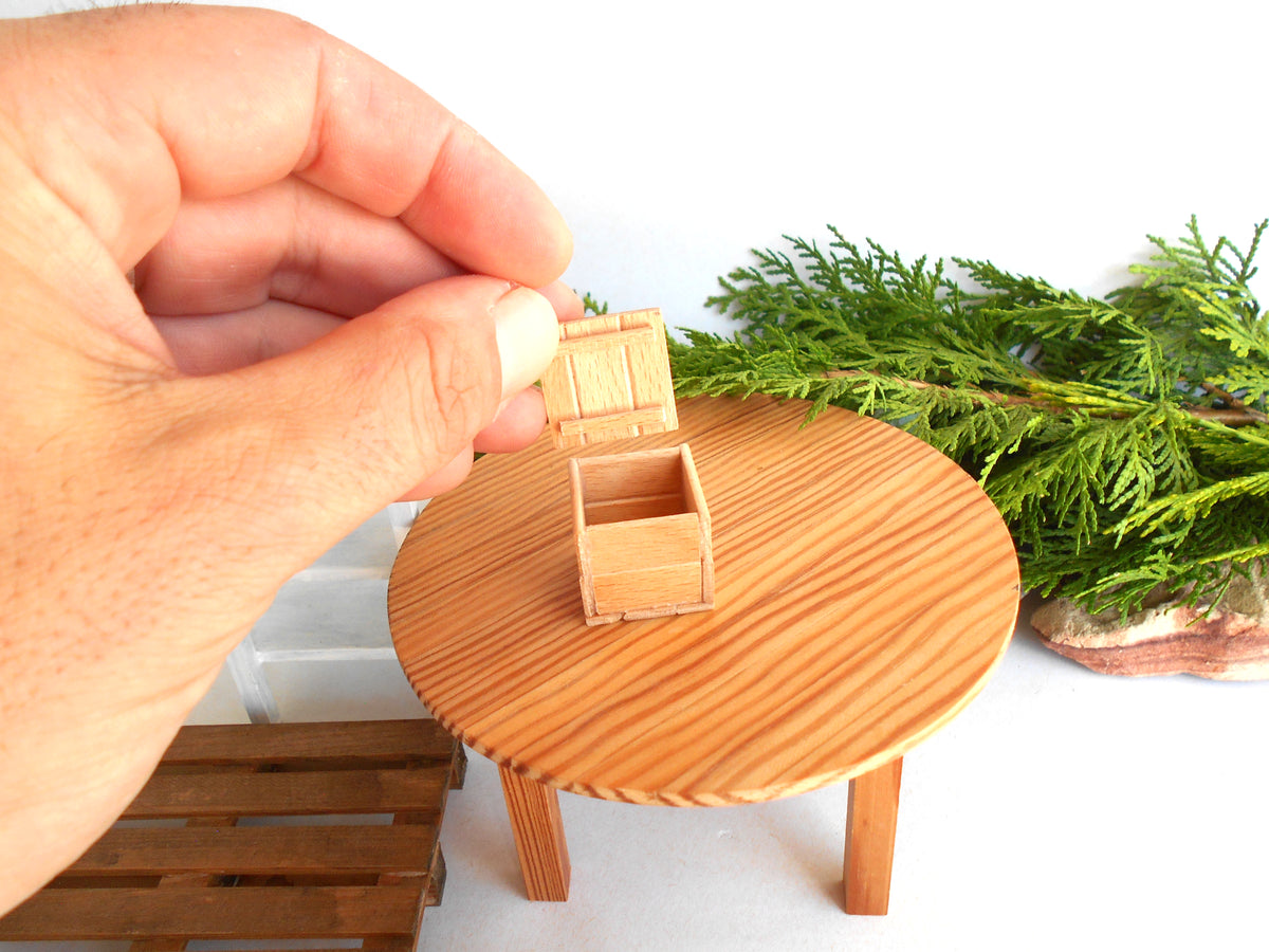 A miniature wooden chest with its lid open, displayed on a small round wooden table. A human hand is holding the chest, and green foliage is visible in the background against a white backdrop.