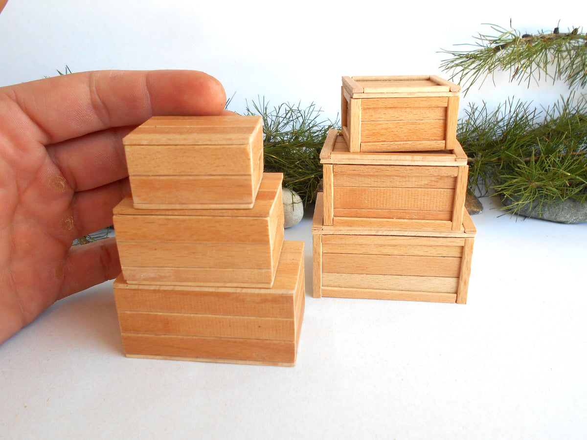 A hand holds a stack of three small natural wood boxes next to a set of two matching wooden boxes on a white surface with green pine branches in the background.