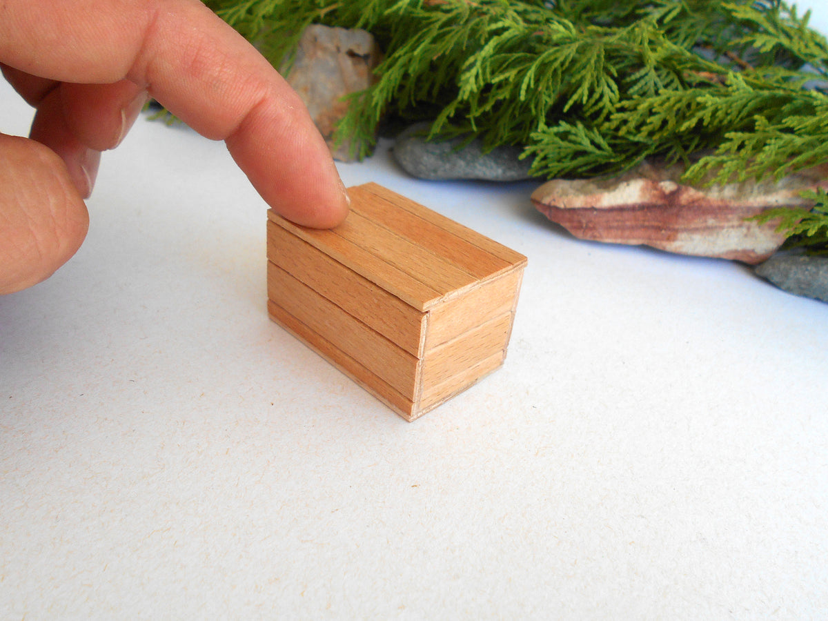 A miniature light brown wooden box with a lid, shown on a white surface with a person's finger pointing at it and green foliage in the background.