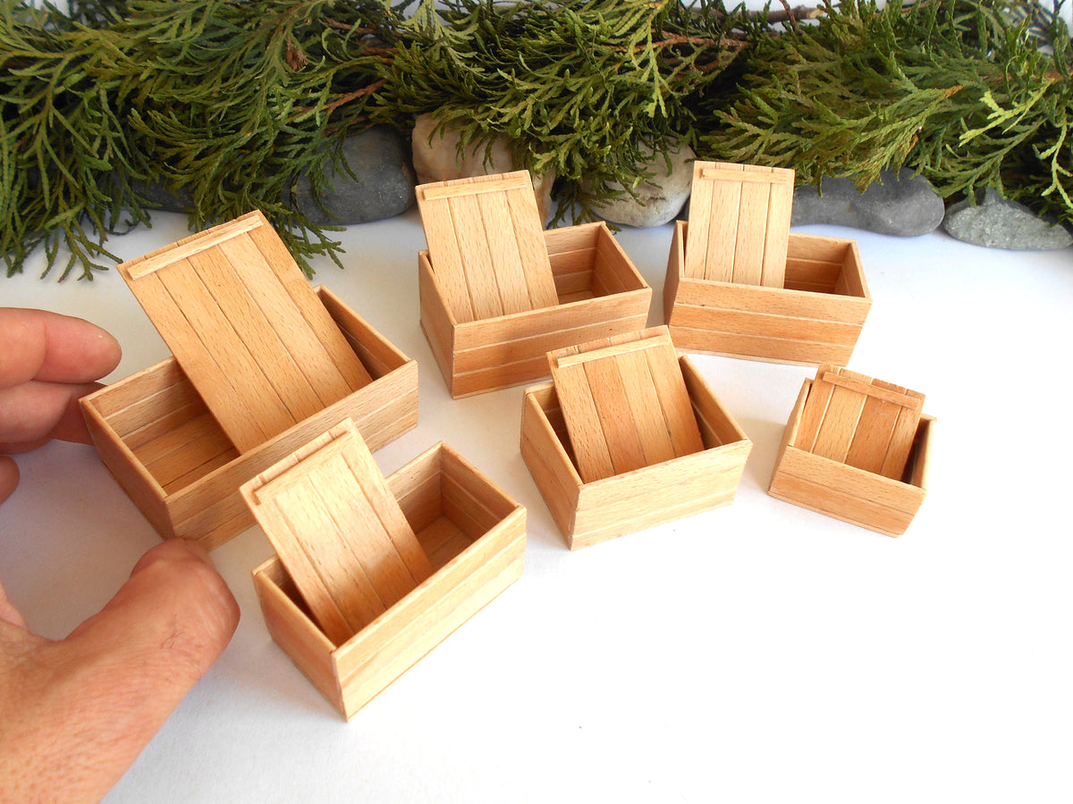 A set of miniature light wood boxes with hinged lids, shown on a white surface with a hand holding one box for scale, and green foliage in the background.