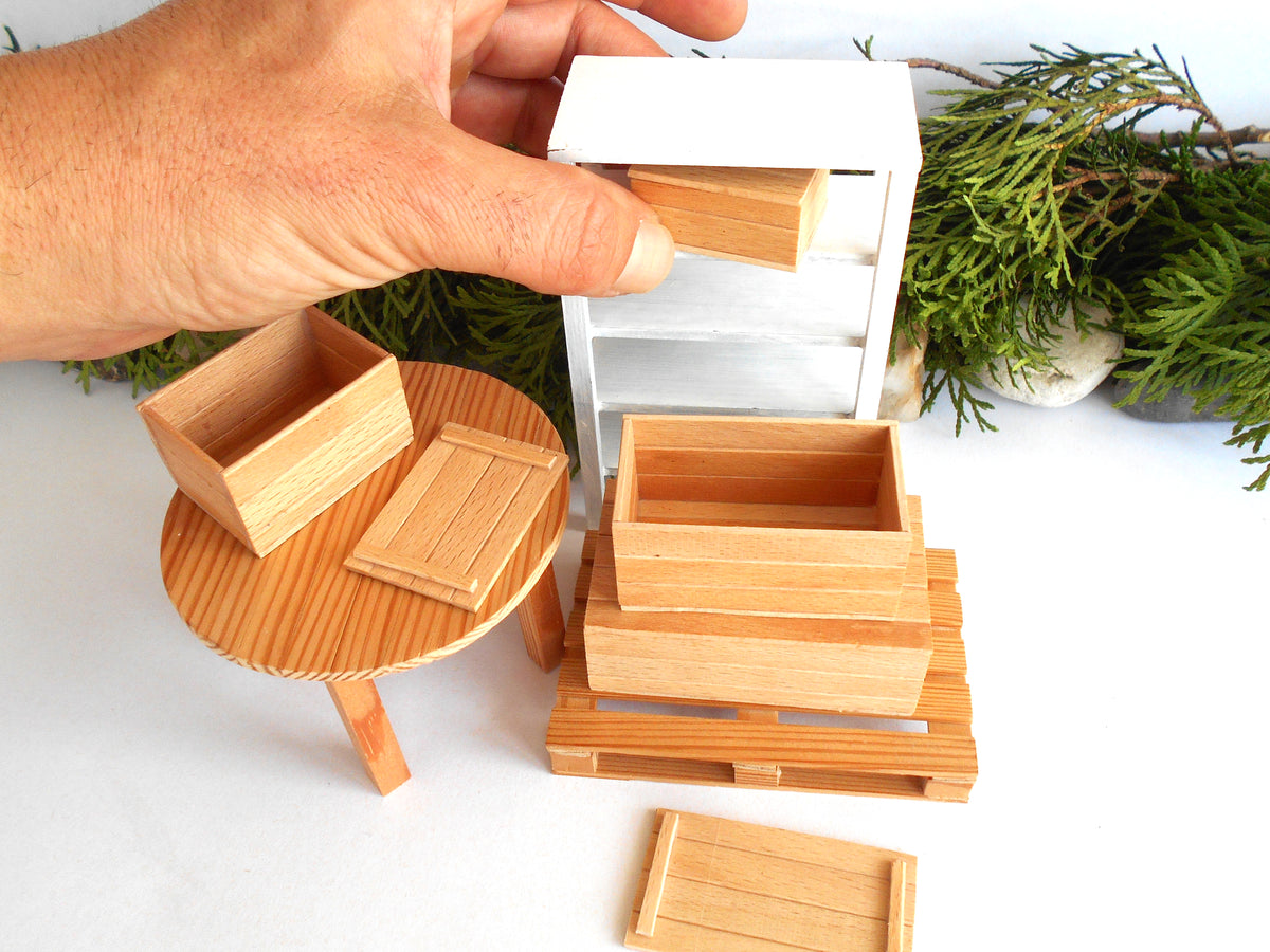 A hand is shown assembling a miniature wooden dollhouse furniture set, including a small white cabinet, a round table, and various wooden crates and pallets, with green foliage in the background.