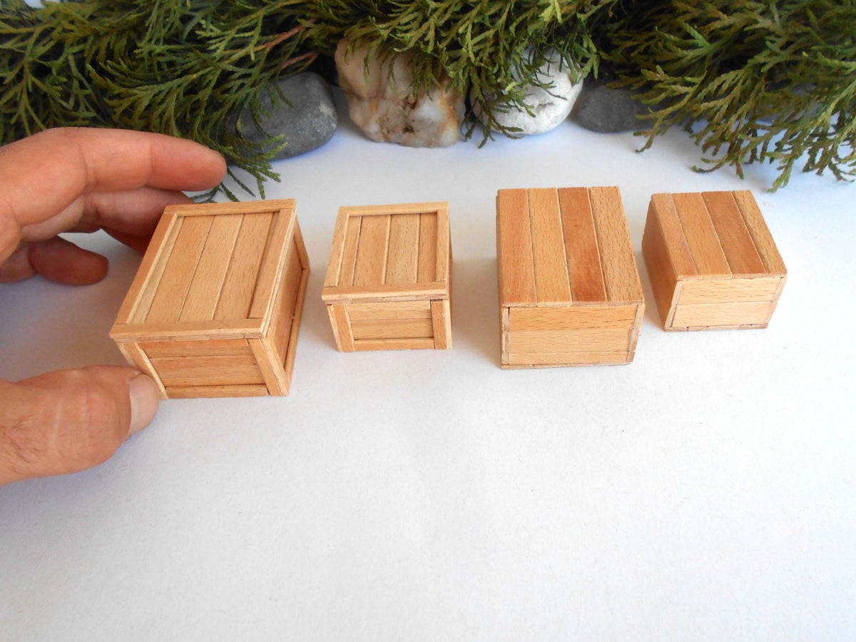Four miniature light wood crates of varying sizes arranged on a white surface, with a hand holding one crate on the left and evergreen branches and stones in the background.