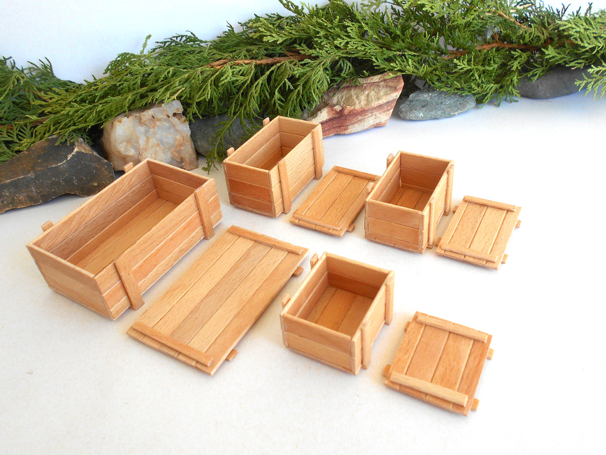 A set of miniature wooden crates and boxes in a light natural wood finish, arranged on a white surface with decorative green pine branches and stones in the background.