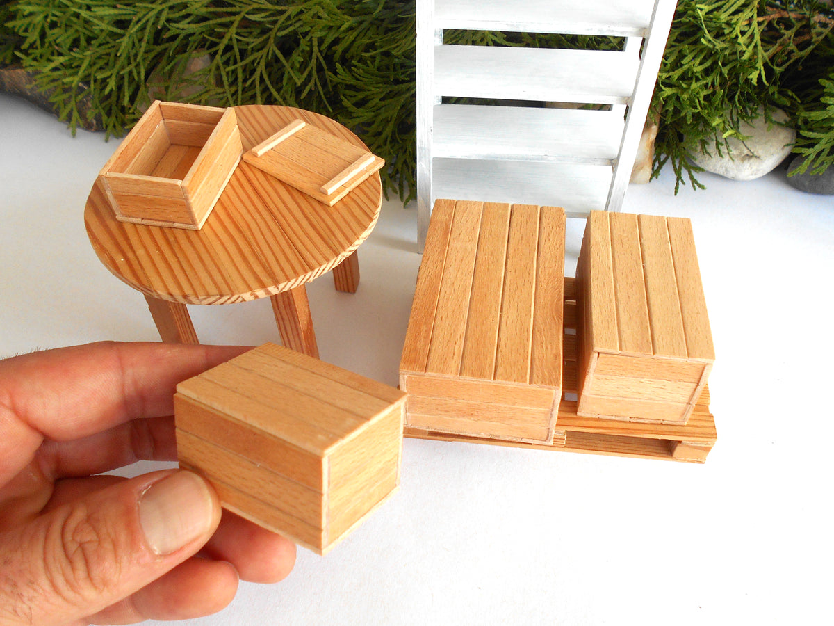 A hand holds a small wooden block in front of a miniature furniture set, including a round table with a box, two tall rectangular boxes on a pallet, and a white slatted chair, all in a light natural wood finish.