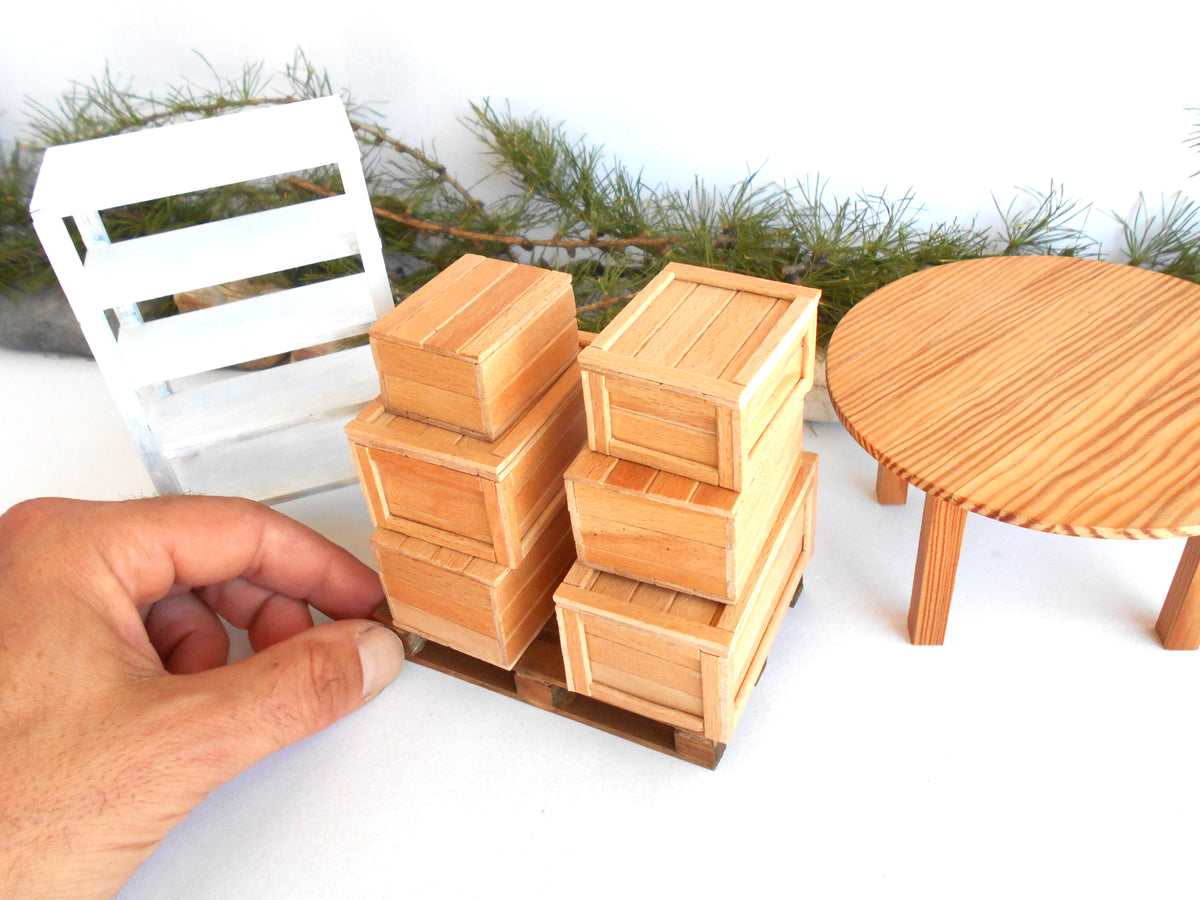 A hand is holding a miniature wooden crate set, featuring two small wooden crates stacked on a miniature pallet next to a miniature white crate and a round wooden table, all set against a white background with pine branches.