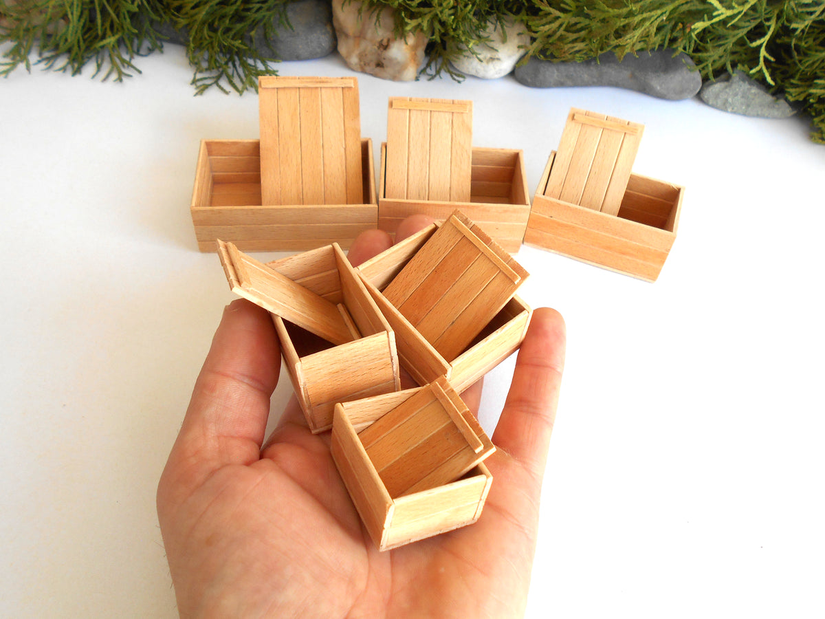 A hand holding several miniature light wood crates, with more of the same crates arranged on a white surface in the background. Small greenery and stones are visible at the top edge.