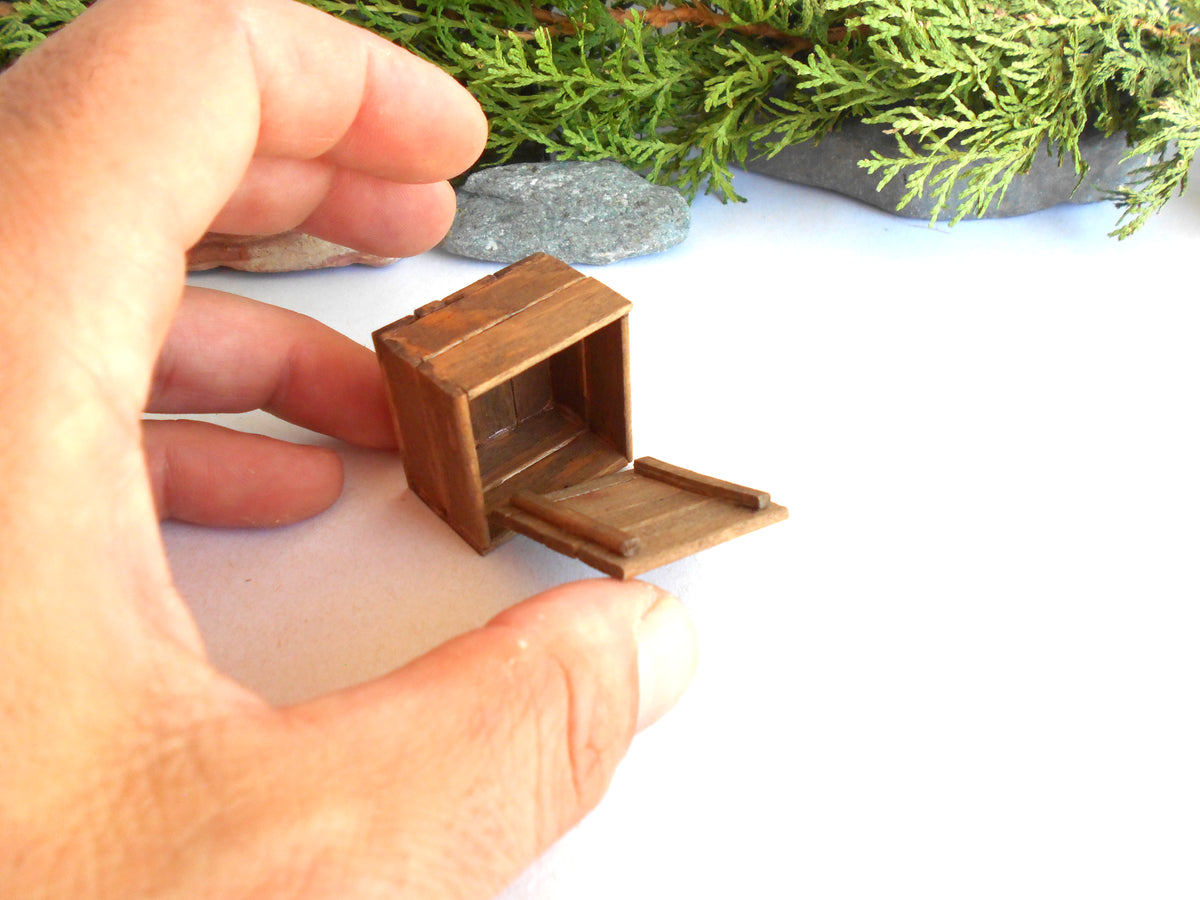 A miniature brown wooden cabinet or storage box with an open door, held by a person's hand against a white background with green foliage and small stones in the upper background.