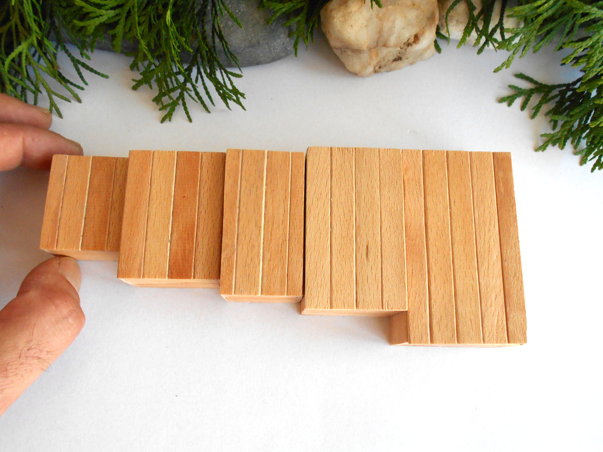A close-up of a hand arranging light natural wood rectangular blocks into a stepped, stair-like structure on a white surface, with greenery and a stone visible in the background.
