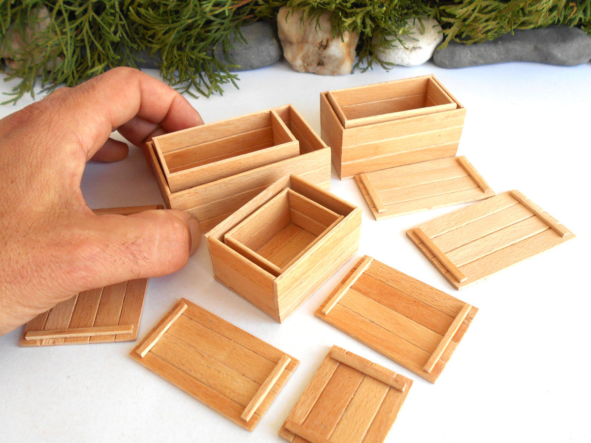 A hand is arranging several small light wood boxes and matching lids on a white surface, with greenery and stones in the background.
