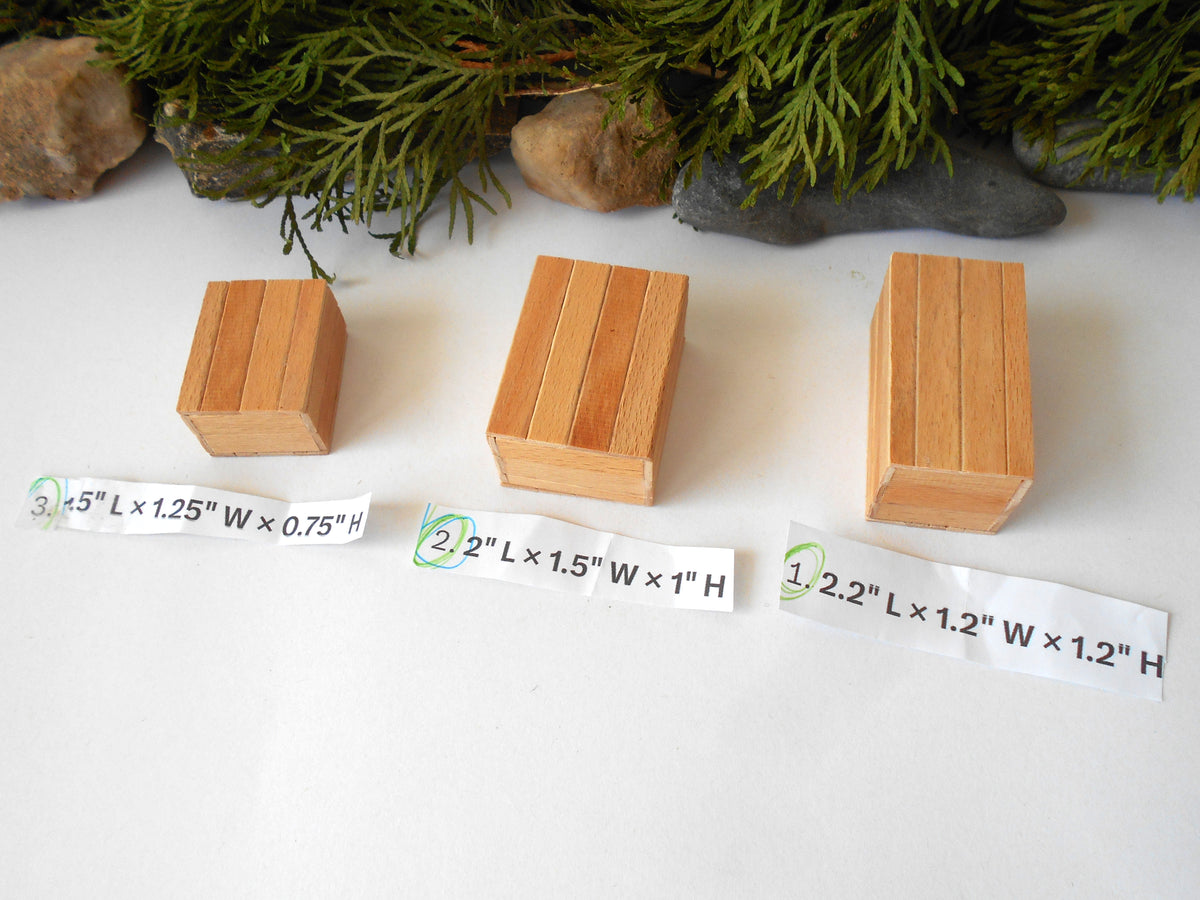 Three small rectangular wooden blocks of different heights arranged on a white surface, with printed size labels underneath each block and evergreen branches and stones at the top.