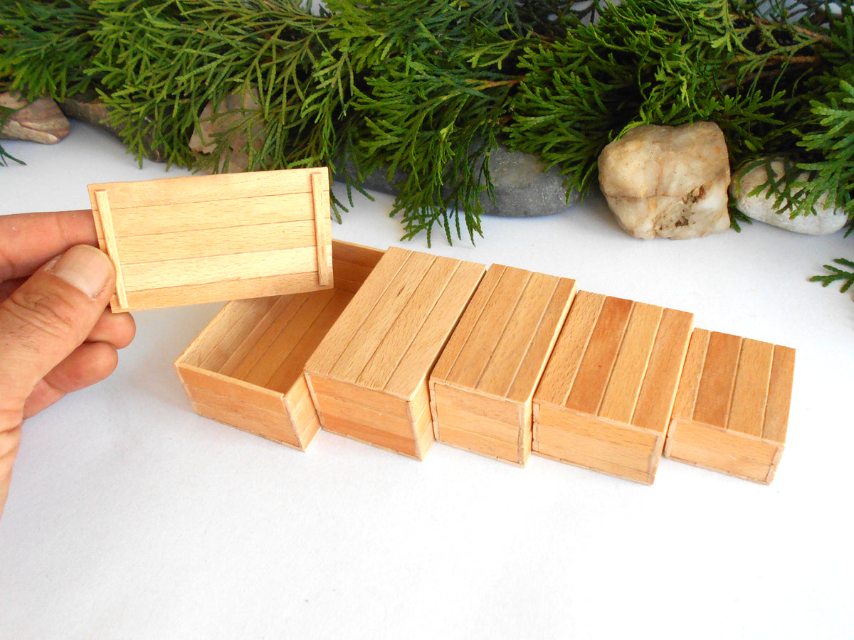A hand holds a small rectangular wooden box lid next to a row of matching small wooden boxes on a white surface, with green foliage and a stone in the background.