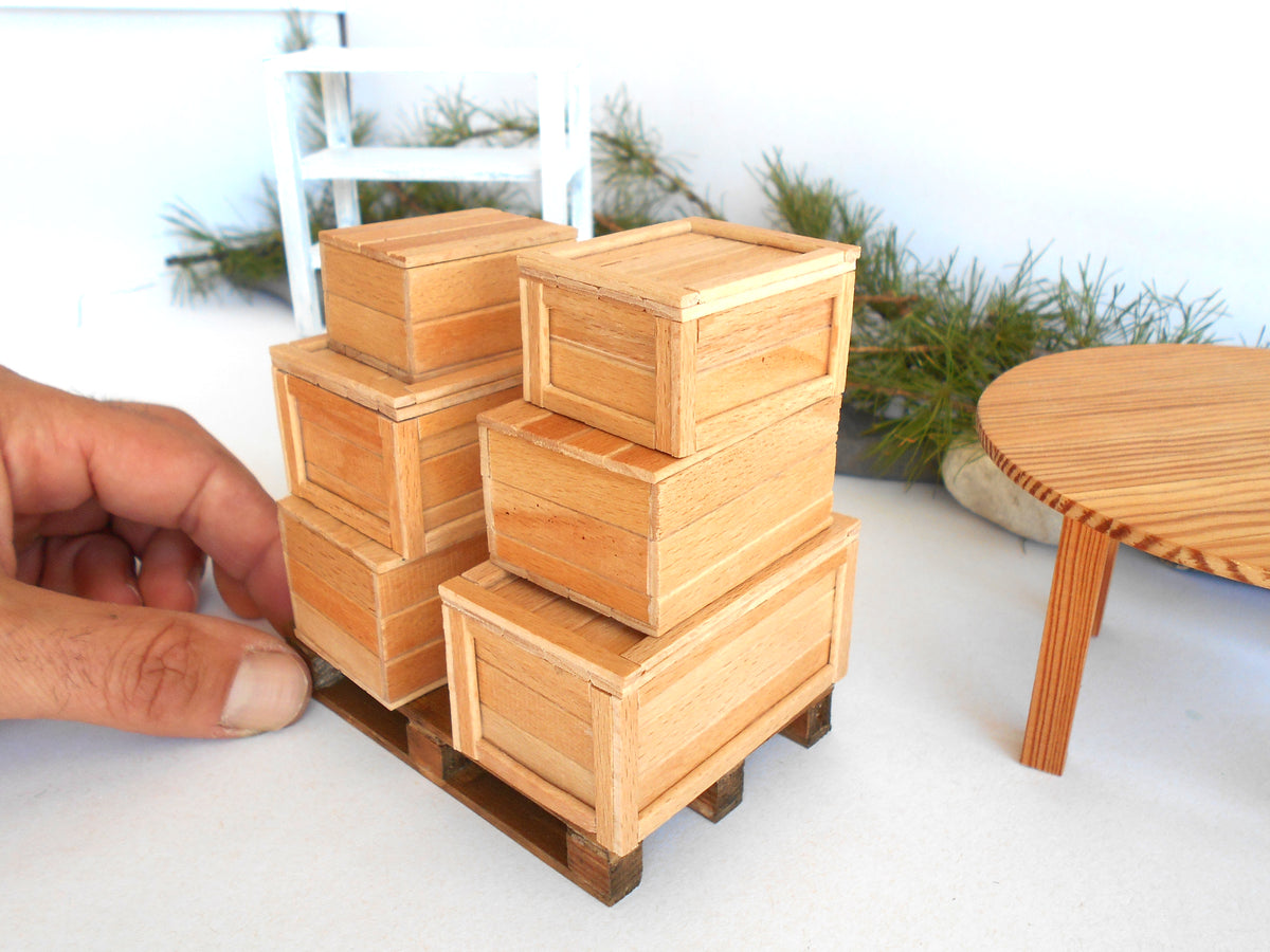 A hand is holding a miniature stack of four small wooden crates on a pallet, likely for a model or diorama. The background features a miniature round table and greenery against a white wall.