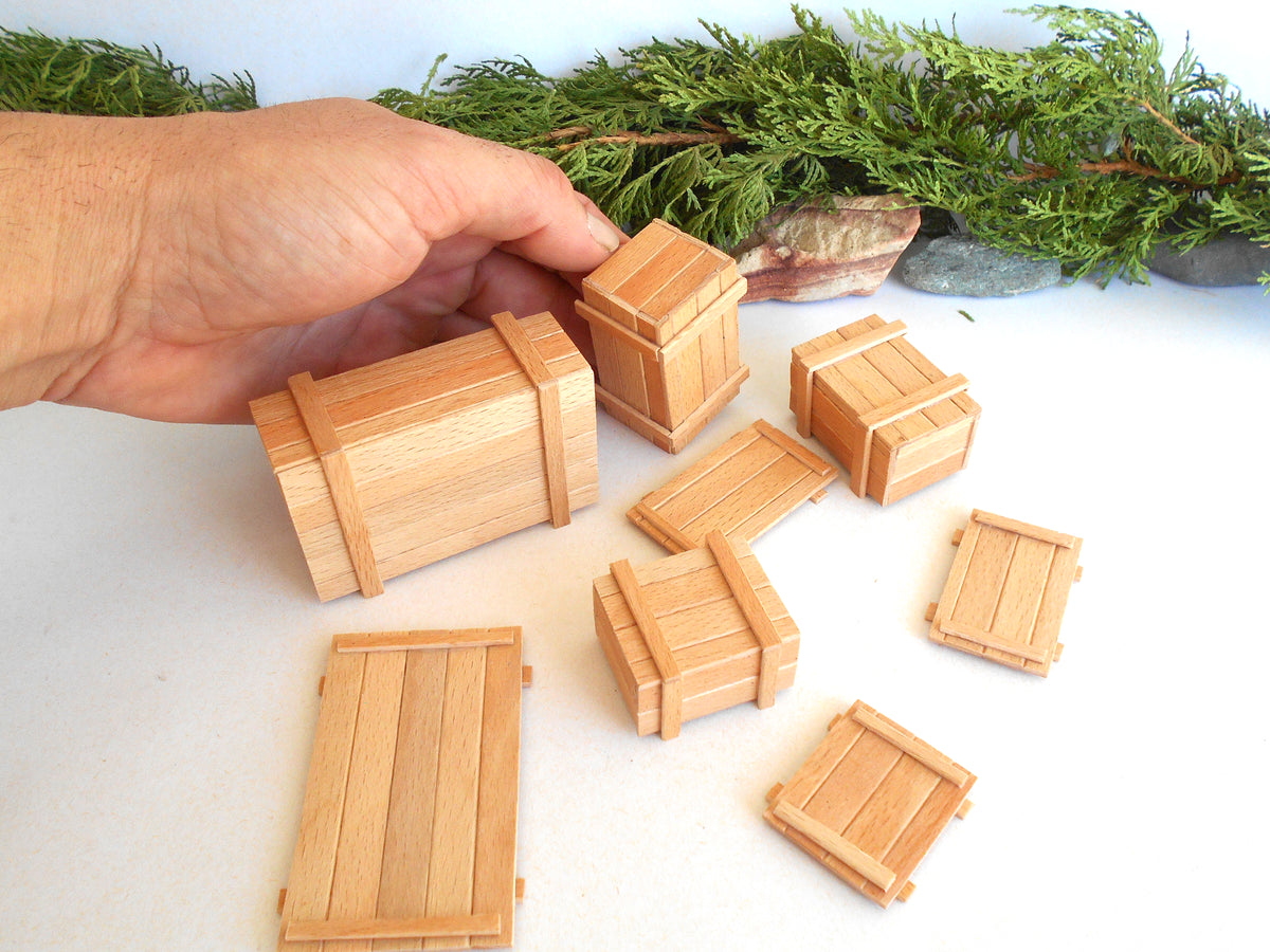 A hand is holding a miniature wooden crate model, with several other small wooden crate and box models scattered on a white surface below, with green foliage and stones in the background.