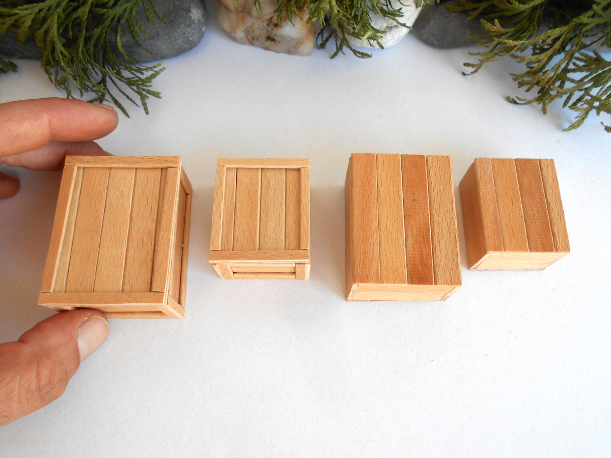 Four miniature wooden crates of varying sizes arranged on a white surface, with a hand holding one of the crates on the left. Small green plants and gray stones are visible in the background.
