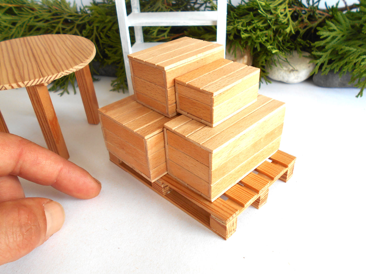 A miniature wooden pallet with stacked wooden boxes, shown next to a miniature wooden table and a person's hand for scale, with artificial green plants in the background.
